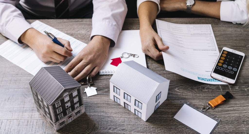 Two individuals exchanging documents over a table, with small model houses and a mobile phone placed on the table, symbolizing insurance solutions for property and financial security.